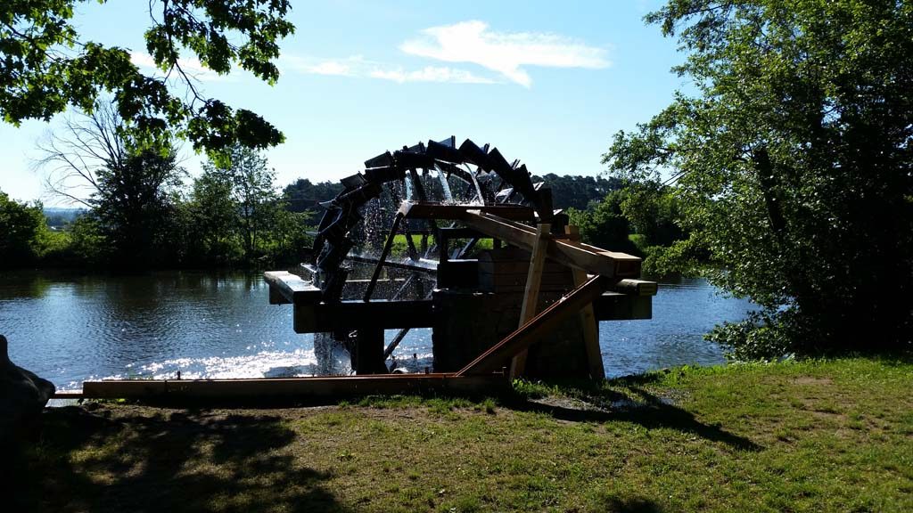 Wasserrad an einem ruhigen Gewässer, umgeben von Bäumen und Sträuchern.